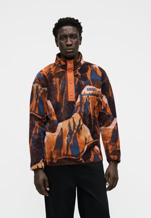 Hombre que lleva un forro polar con estampado multicolor en naranja, azul y negro con botones automáticos naranjas y pantalones negros, de pie frente a un fondo liso.