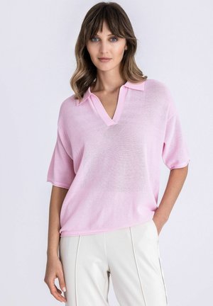 Frau mit welligem braunem Haar, die ein locker sitzendes hellrosa kurzärmeliges Hemd mit Kragen und weiße, hoch taillierte Hose trägt, steht mit einer Hand in der Tasche.