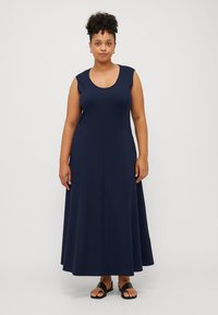 Mörkblå ärmlös maxiklänning i mjukt tyg, med rund halsringning, utsvängd silhuett och sömlös design. Bärs med svarta sandaler.