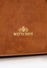 Sac en daim marron arborant un logo doré "WITTCHEN" avec des détails de couronne et de blason de lion élaborés. Texture lisse avec un léger éclat.