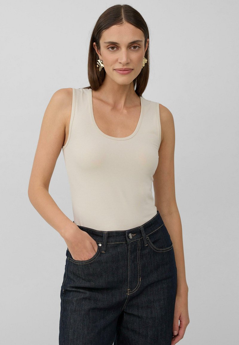 Top beige senza maniche con scollatura a barca, realizzato in tessuto morbido. Abbinato a jeans in denim scuro con un classico design a cinque tasche.