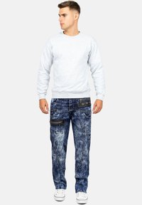 Felpa grigia abbinata a jeans cargo blu con effetto usurato, dotati di tasche con zip e un lavaggio chiaro. Gli accessori includono sneakers bianche.