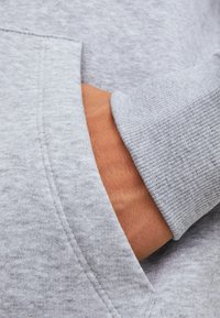 Gray sweatshirt fabric with a soft texture, featuring a kangaroo pocket. Close-up of a hand resting in the pocket, showing skin tone and detail.