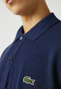 Polo shirt navy en tissu texturé, avec un col classique, deux boutons, et un logo de crocodile vert brodé sur la poitrine gauche.