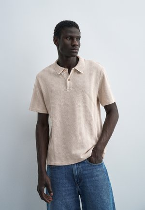 Schwarzer Mann steht, trägt ein beiges kurzärmeliges Poloshirt mit Chevron-Muster und blaue Jeans, vor einem einfarbigen hellgrauen Hintergrund.