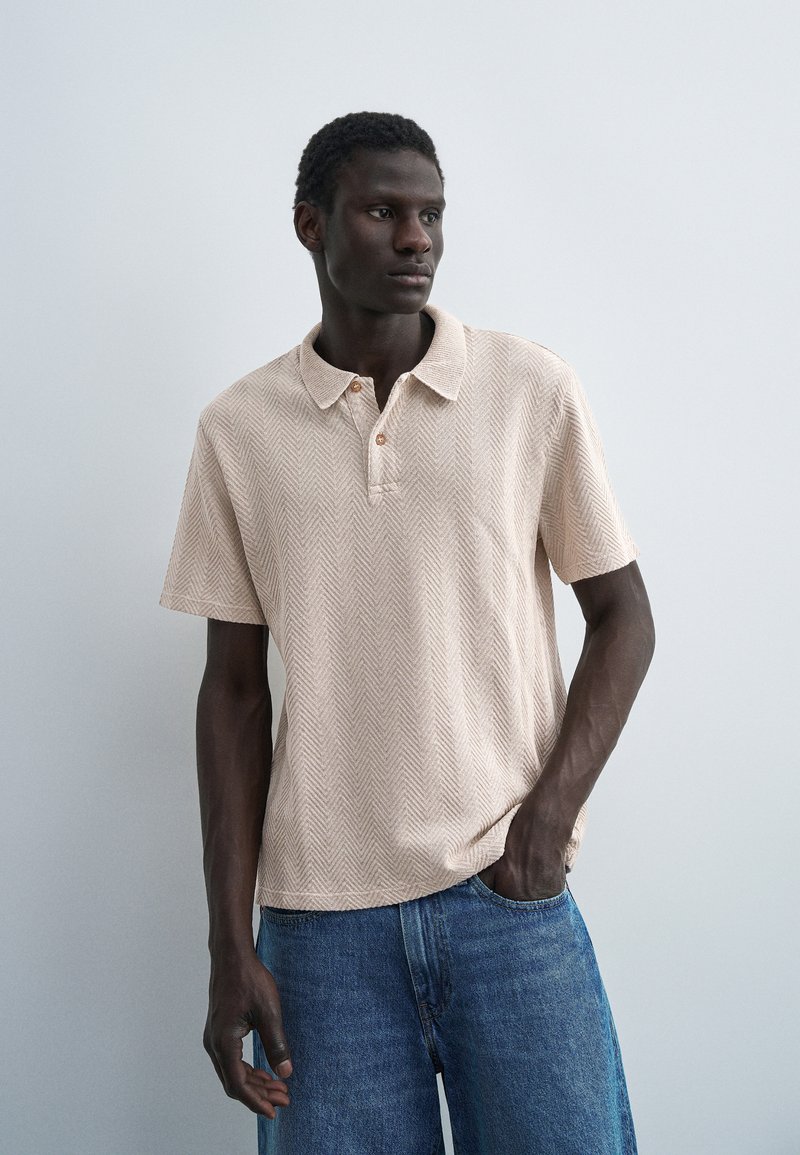 Μαύρος άντρας στέκεται, φοράει μπεζ κοντομάνικη πόλο με σχέδιο ζιγκ-ζαγκ και μπλε τζιν παντελόνι, πάνω σε ένα μονόχρωμο ανοιχτό γκρι φόντο.