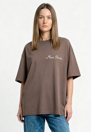 Giovane donna con capelli castani lisci indossa una t-shirt marrone oversize con la scritta "Mira Paris" e jeans blu su uno sfondo chiaro e semplice.
