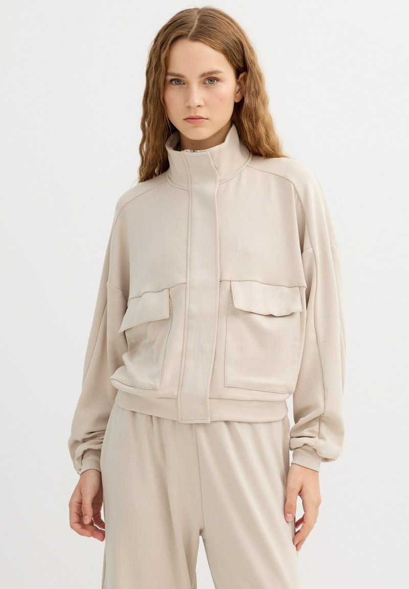 Chaqueta corta de color beige claro hecha de tela suave, con cuello alto, dos bolsillos delanteros y un detalle de costura en contraste.