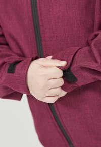 Enfant ajustant la sangle noire en Velcro sur la manche d'une veste zippée bordeaux.