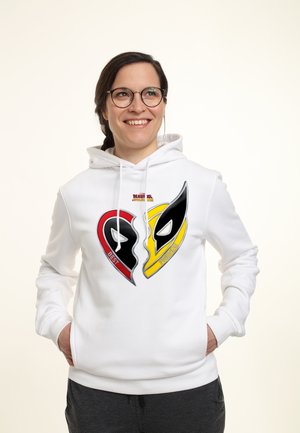 Uśmiechnięta kobieta w okularach ubrana w biały kaptur z grafiką złamanego serca w czerwonym i żółtym kolorze z maskami Deadpoola i Wolverinea.