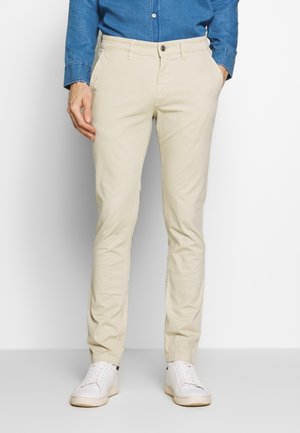 Mann trägt beige, eng anliegende Hose, ein blaues Jeanshemd, weiße Turnschuhe, steht mit einer Hand in der Tasche vor einem schlichten Hintergrund.