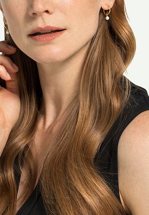 Goldene Creolen mit einem kleinen weißen Perlenakzent, vor einem Hintergrund aus langen, gewellten kastanienbraunen Haaren und einem schwarzen Top.