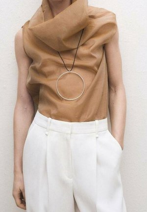 Osoba nosząca beżowy top bez rękawów z drapowanym materiałem i dużym okrągłym naszyjnikiem, w połączeniu z białymi, wysokim stanem spodnimi.