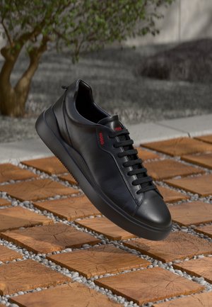 Czarny skórzany sneaker z czarnymi sznurowadłami i czerwonym logo "HUGO", unoszący się nad drewnianą podłogą z przerwami wypełnionymi żwirem.