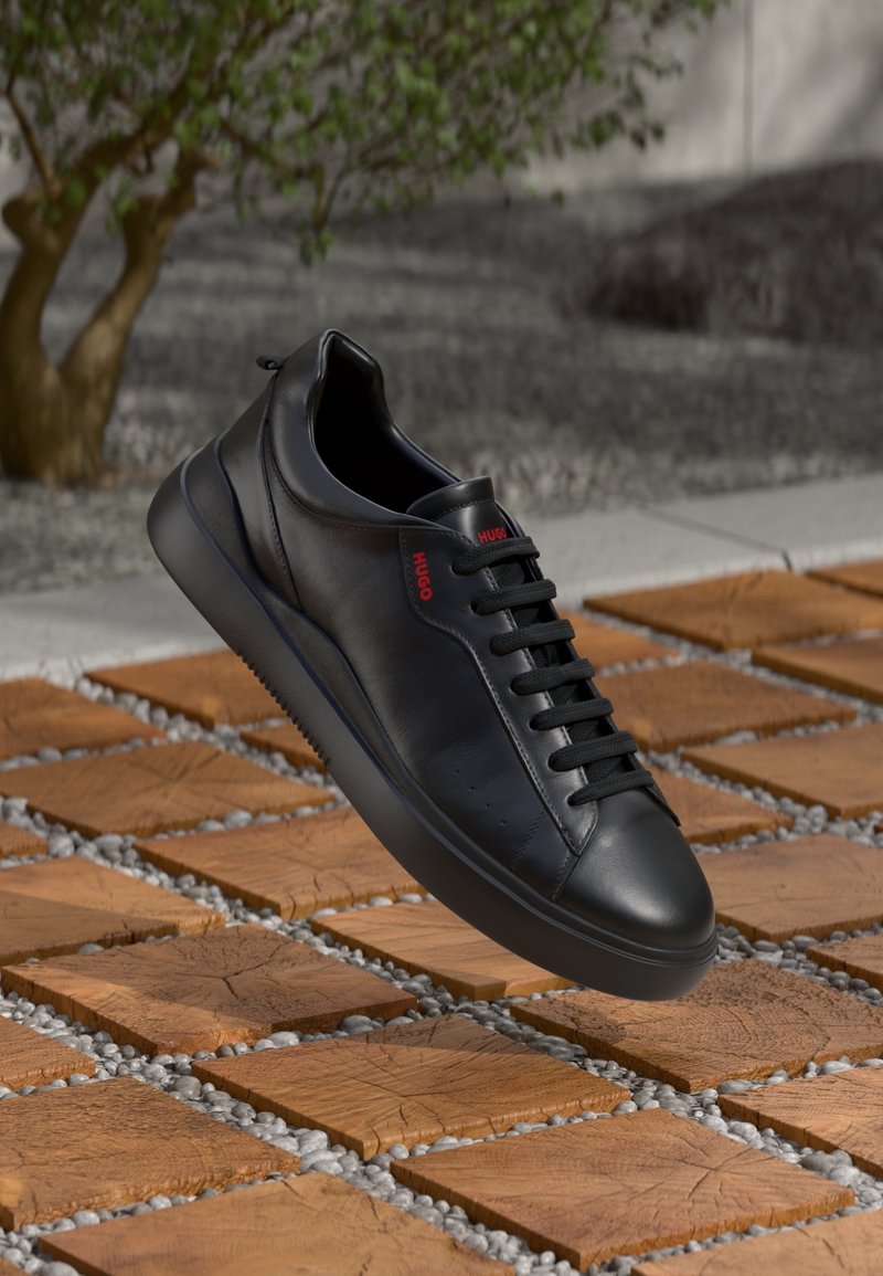 Baskets en cuir noir avec lacets noirs et logo rouge "HUGO", flottant au-dessus d'un sol de carrelage en bois avec des espaces remplis de gravier.