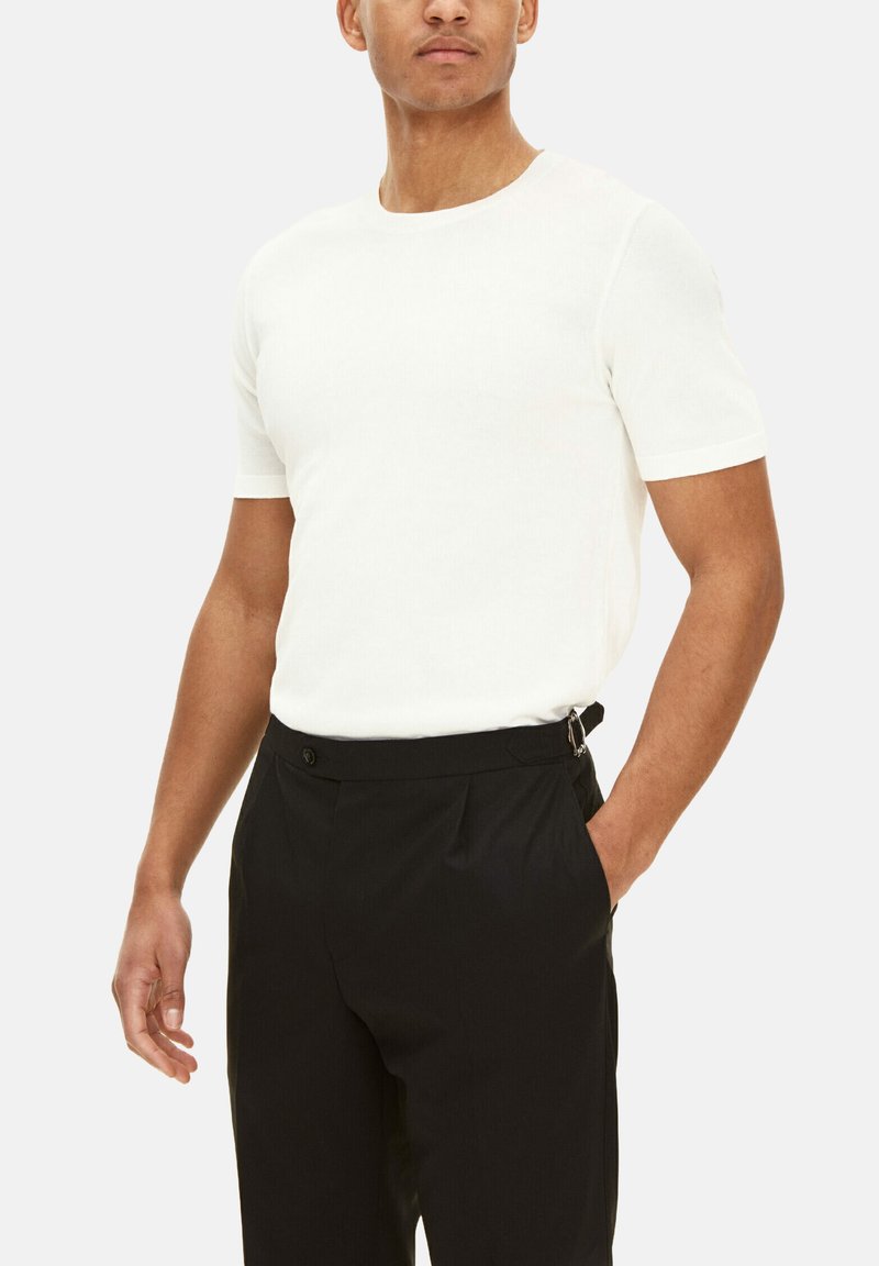 Camisetta bianca a maniche corte con texture a coste, indossata con pantaloni neri che presentano tasche laterali e un dettaglio con chiusura in vita.