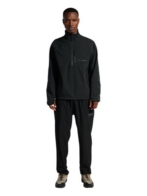 Vyras vilki juodą užtrauktuku užsegamą švarką ir juodas kelnes su pilkais ir juodais sportbačiais, stovintis prieš baltą foną.