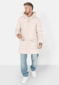 Lichtbeige pufferjas met een capuchon, voorzien van twee voorzakken, een quilted ontwerp en verstelbare manchetten, gedragen over blauwe jeans en witte sneakers.