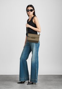 Olíva zöld keresztpántos táska, fedélzáras, sima textúrával és minimális fém részletekkel. Széles szárú kék farmerral és fekete ujjatlan topal viselve.