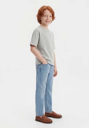 Rødthåret dreng smilende, iført en lysegrå kortærmet skjorte, lyseblå jeans, brune loafers, stående med den ene hånd i lommen.