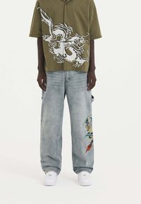 Olivgrön kortärmad skjorta med vit drakentryck; ljusblå baggy jeans med färgglödig brodyrdesign på benet; vita sneakers.