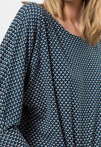 Bluse mit blauem Muster, das ein geometrisches Design mit quadratischen und Pfeilmotiven zeigt, aus einem leichten Stoff mit glatter Textur gefertigt.