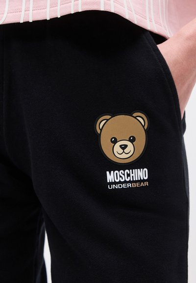 Άτομο που φοράει μαύρο παντελόνι με λογότυπο καφέ αρκούδας και κείμενο «Moschino Underbear» κοντά στην τσέπη, με το χέρι μέρος μέσα στην τσέπη.