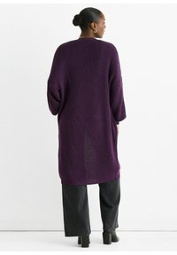 Purple knitted cardigan with a loose, oversized fit and long sleeves. Features a textured pattern and side slits. Paired with dark jeans and block-heel shoes.