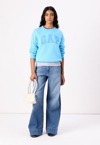 Sudadera de algodón azul claro con el logo "GAP", combinada con unos pantalones vaqueros de pierna ancha en azul desgastado. La modelo sostiene un pequeño bolso blanco. Fondo liso.