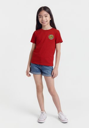 Meisje met lang donker haar, gekleed in een rood Santa Cruz T-shirt, spijkershorts en witte sneakers, staand en glimlachend tegen een witte achtergrond.