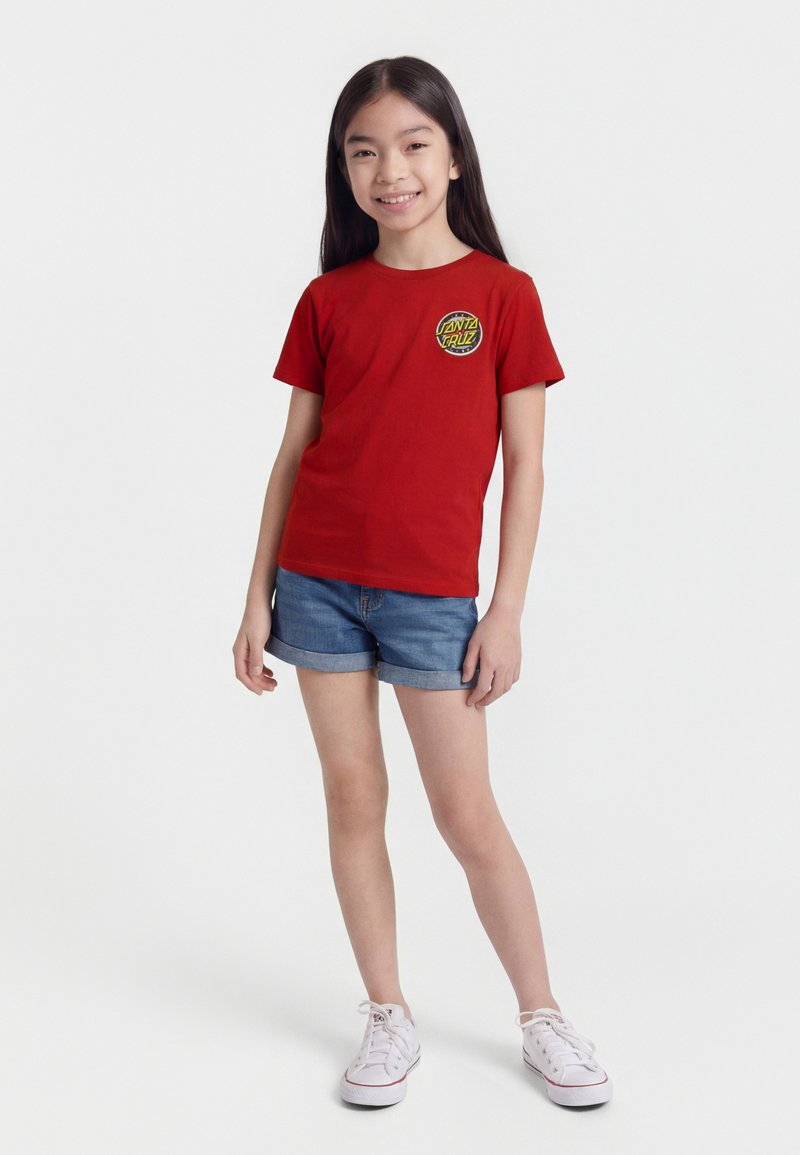 Mädchen mit langen dunklen Haaren, das ein rotes Santa Cruz T-Shirt, Jeansshorts und weiße Sneaker trägt, steht lächelnd vor einem weißen Hintergrund.