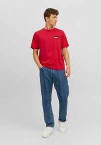 Rode katoenen t-shirt met een klein zwart-wit logo op de borst, gecombineerd met lichtblauwe spijkerbroeken en witte sneakers.