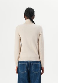 Beige høyhalset genser med lange ermer, rillede mansjetter og kant, sammen med løstsittende blå denimjeans, sett fra baksiden.