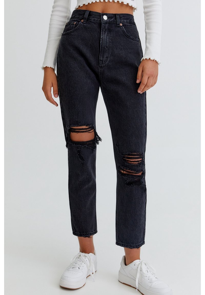 Black high-waisted jeans with distressed rips, featuring a five-pocket design and metal button closure, paired with white sneakers.