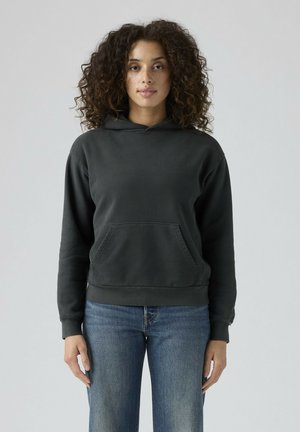 Vrouw met krullend haar, gekleed in een donkergrijze hoodie en een blauwe spijkerbroek, staand tegen een effen lichte achtergrond.