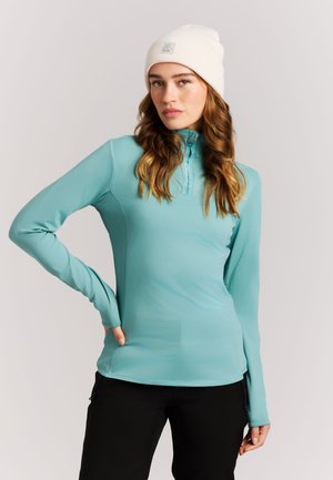 Femme portant un bonnet blanc et un haut à manches longues bleu clair avec un pantalon noir, debout avec une main sur la hanche devant un fond uni.