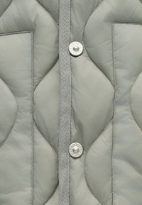 Κουβερλί μπουφάν σε ανοιχτό γκρι χρώμα, με υφή, δύο μπροστινές τσέπες και ασημένια κουμπιά σνίκερ κατά μήκος της κεντρικής καπάκης.