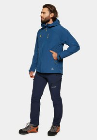 Niebieska kurtka outdoorowa z zamkiem błyskawicznym z przodu, kapturem i teksturowanym materiałem. Połączona z granatowymi spodniami z bocznymi kieszeniami i szarymi butami trekkingowymi.