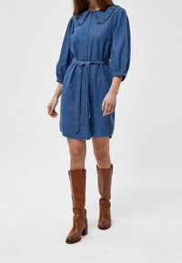 Jeansklänning med volangkrage, midjebälte och ballongärmar, matchad med bruna läderstövlar i knähöjd. Ljusblå färg, mjuk textur.