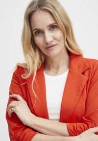 Blonďatá žena s vlnitými vlasy, oblečená v oranžovém saku přes bílé tričko, se zkříženýma rukama, klidně se dívající do kamery na bílém pozadí.
