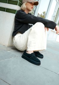 Sorte platformssko i ruskind med en glat tekstur, rundet snude og snørelukning, kombineret med cremegule bukser med brede ben og en mørk sweater.