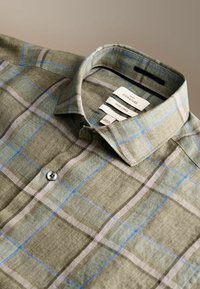 Světlá olivově zelená košile z lněné látky s modrými a béžovými pruhy, zapnutým límcem a viditelnými značkami značky a látky uvnitř.