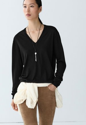 Donna che indossa un maglione nero a scollo a V, pantaloni beige, una giacca bianca legata intorno alla vita e una lunga collana con un ciondolo a forma di chiave.