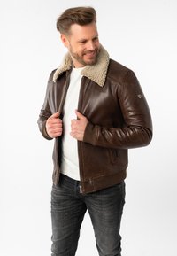 Giacca di pelle marrone con colletto in shearling, chiusura con zip, polsini a coste e tasche laterali. Indossata sopra una camicia bianca e abbinata a jeans scuri.