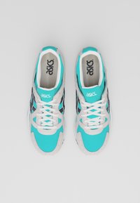 Ett par turkosa och grå Asics sneakers med vita snören, sedd ovanifrån på en enfärgad ljusgrå bakgrund.