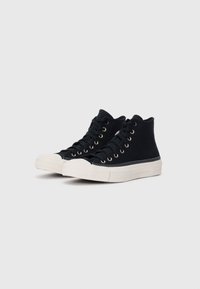 Sneaker alte in tela nera con occhielli dorati, punta in gomma bianca e suola testurizzata. Progettate per un uso casual con un'estetica minimalista.