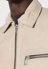 Beige Lederjacke mit Kragen, ausgestattet mit einem Frontreißverschluss und einer Reißverschlusstasche mit silbernen Beschlägen, glatte Textur und minimalistisches Design.