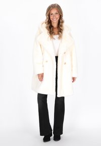 Kvinde, der smiler, iført en hvid faux fur frakke, hvid top, sorte flared bukser og sorte støvler, stående mod en hvid baggrund.