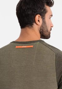 Maglione verde oliva da uomo con tessuto testurizzato, colletto a coste e un'etichetta arancione del marchio sul retro. Presenta un design aderente.