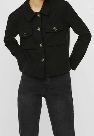 Mujer con chaqueta negra abotonada con dos bolsillos con solapa delanteros, combinada con vaqueros de tiro alto en gris oscuro.